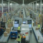 Inside a busy warehousing and fulfilment centre with workers handling packages and conveyor belts moving parcels towards delivery trucks.